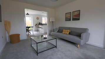 Una sala de estar con una mesa de centro de vidrio, un sofá gris con un cojín estampado, una lámpara y dos cuadros colgados en la pared.