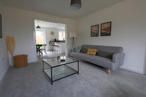 Un salon avec une table basse en verre, un canapé gris agrémenté d’un coussin à motifs, une lampe et deux tableaux accrochés au mur.