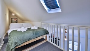 A bedroom with a sloped ceiling, a skylight, a bed with white linens, a green bedspread, a white railing, and a hanging light fixture. - Modern 1-bed Flat Near Cardiff City Centre (Cardiff)