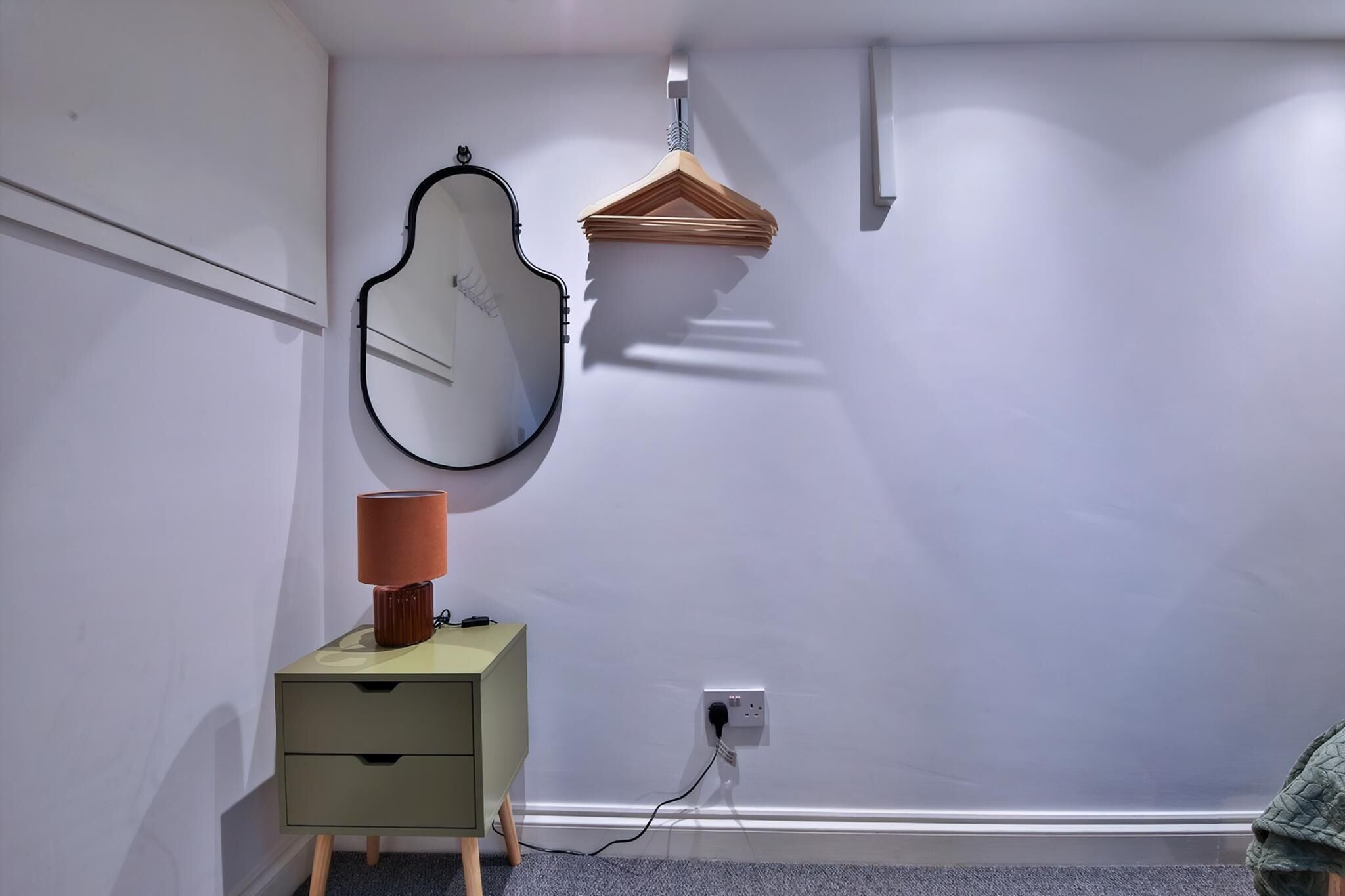 Une chambre avec une seule table de chevet, une lampe, un porte-manteau en bois suspendu, un miroir fixé au mur et une prise de courant.