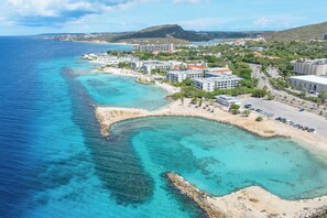 An aerial view of a coastal area with a beach, clear blue waters, and buildings.