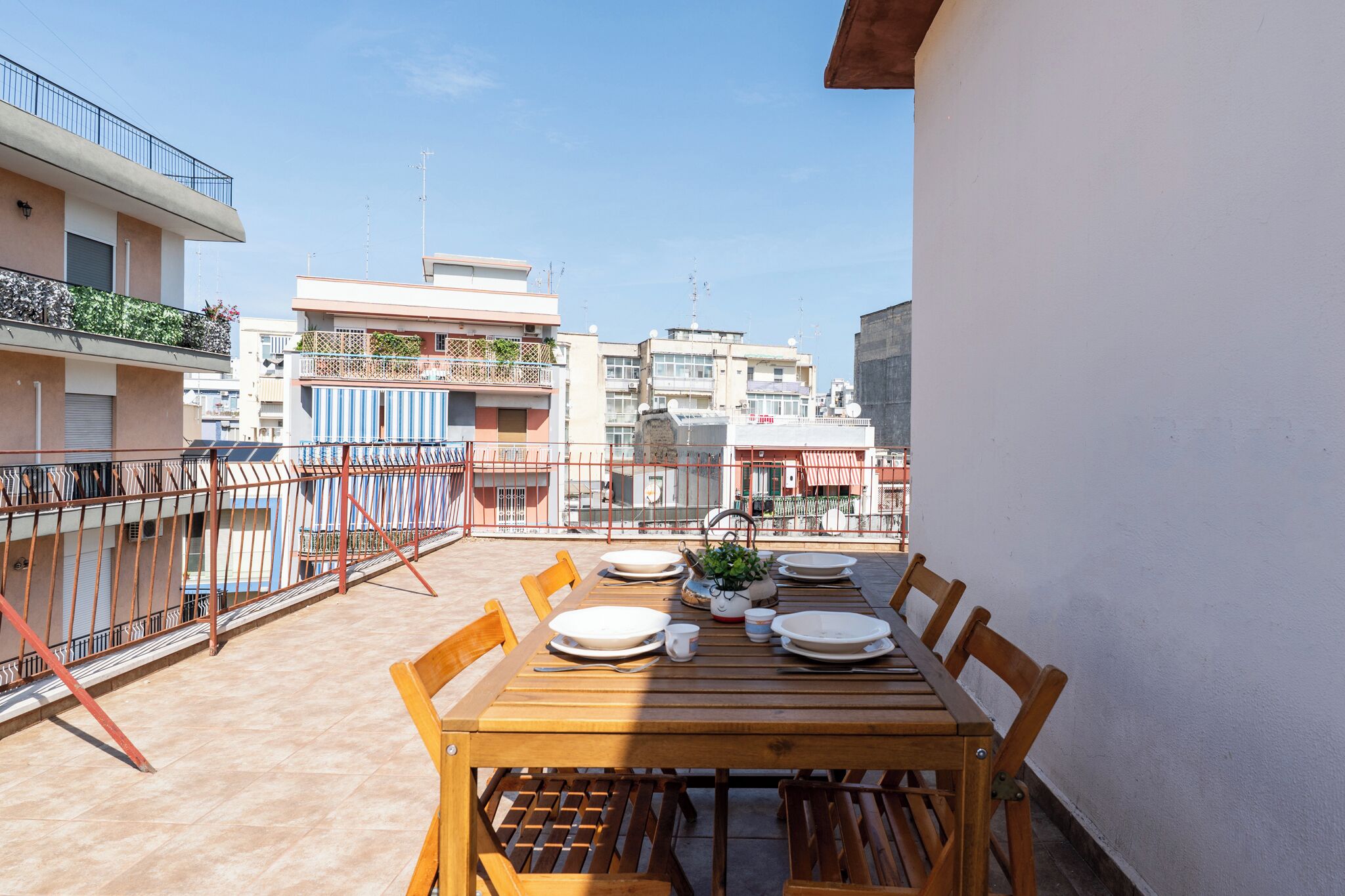 Une table en bois, dressée pour un repas, est placée sur un balcon d’où l’on peut admirer les bâtiments environnants.