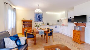 A modern living room with a sofa, dining table, and chairs, a kitchen area with cabinets and appliances, and a TV mounted on the wall. - Holiday Home 'Maison Tournesol' with Shared Pool, Wi-Fi and Air Conditioning (Castelnaud-la-Chapelle)