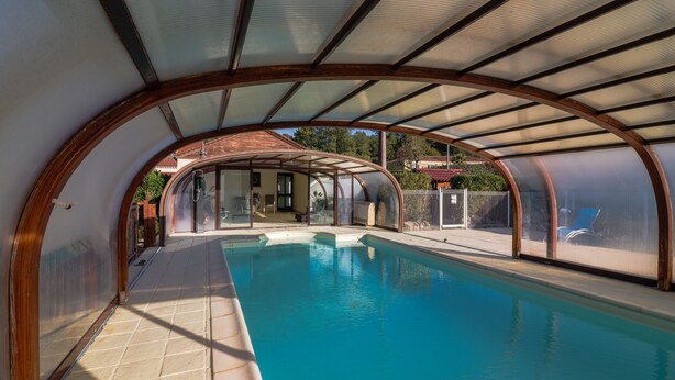 An indoor swimming pool with a curved roof structure, clear glass walls, and a tiled floor. - Holiday Home 'Maison Tournesol' with Shared Pool, Wi-Fi and Air Conditioning (Castelnaud-la-Chapelle)