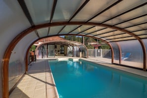 Una piscina interior amb una estructura de teulada corbada, parets de vidre transparent i un paviment de rajola.