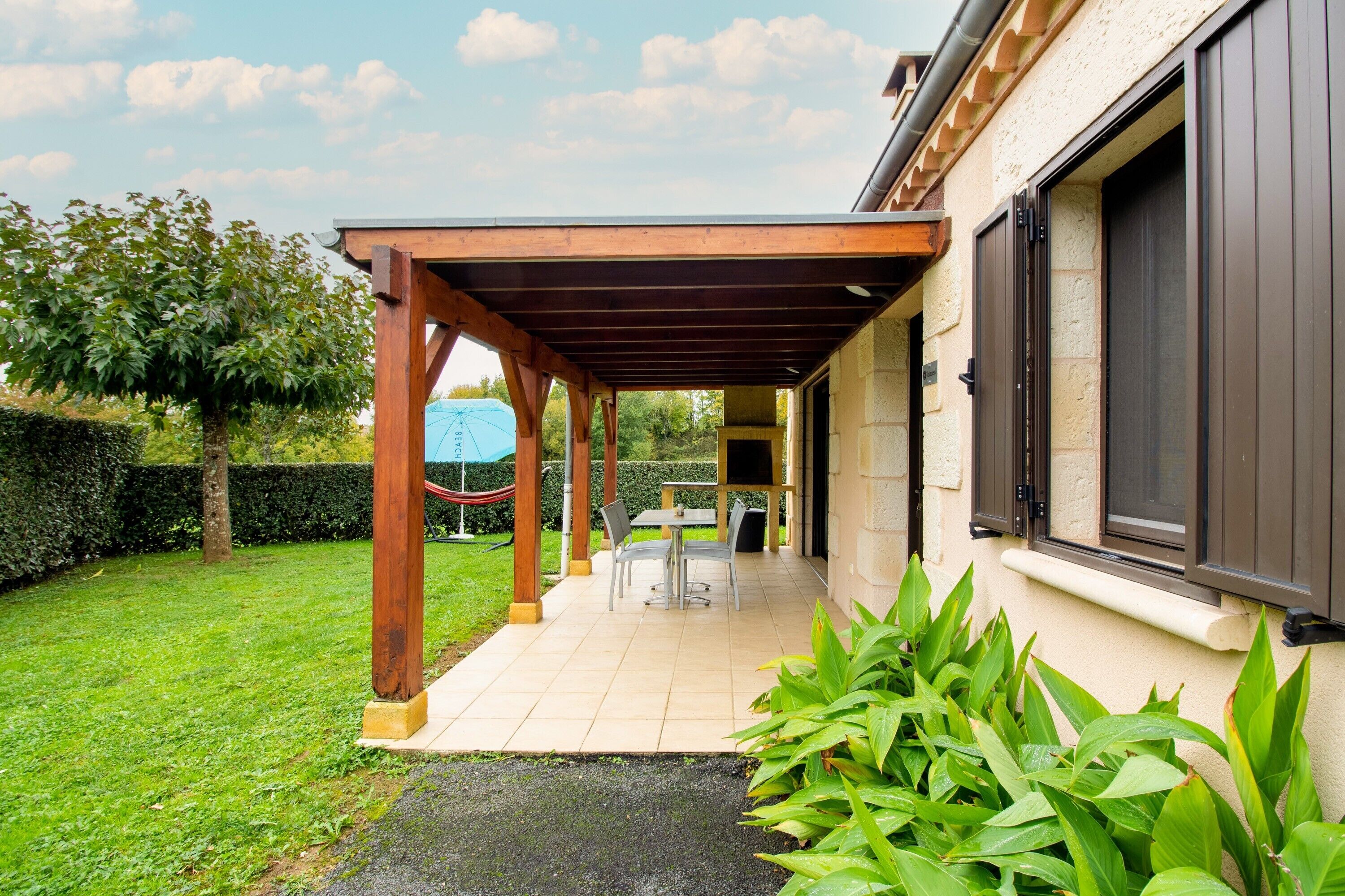 A covered patio area with a table and chairs, a grassy lawn, and a building with a beige exterior and dark shutters.