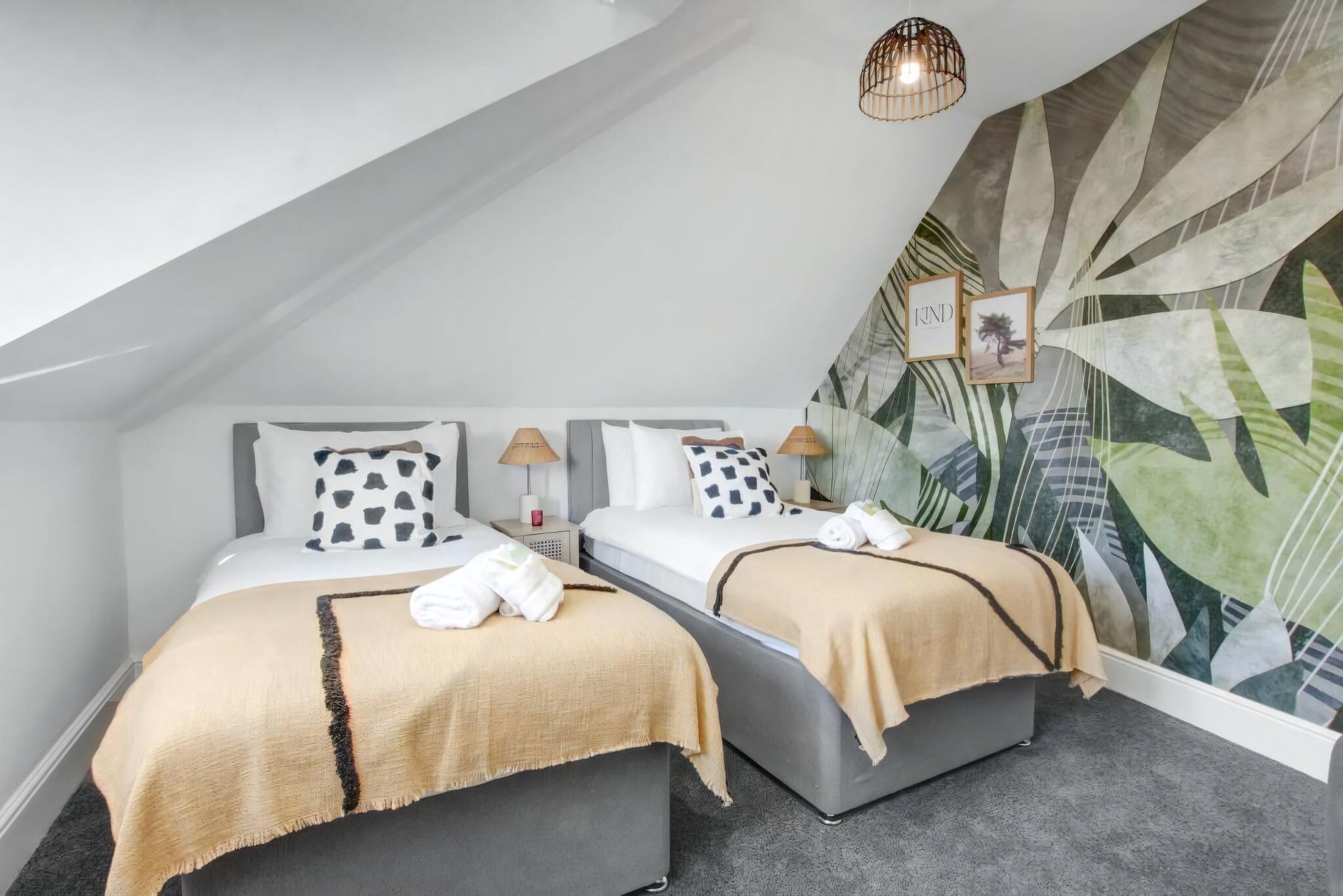 Une chambre d’hôtel moderne avec deux lits, un plafond en pente, un mur décoré de motifs végétaux et un petit luminaire suspendu.