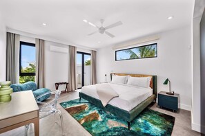 A modern hotel room with a large bed, a desk, and a chair. There is a window with a view of palm trees and a balcony. - Villa Jupiter - Oceanview with Private Pool (Providenciales)