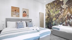 Habitación de hotel con una cama grande, una cama más pequeña, un cabecero gris, dos cuadros colgados en la pared, una puerta y un mural vegetal grande.