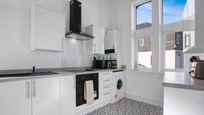 Une cuisine moderne avec des meubles blancs, un four encastré, un lave-linge et une grande fenêtre qui laisse entrer la lumière naturelle.