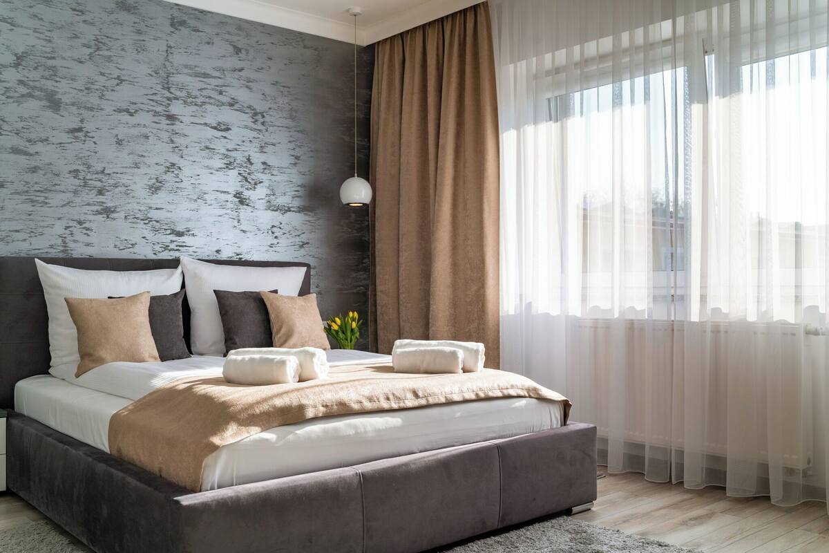 A modern bedroom with a large bed, a grey textured wall, a hanging light, and sheer curtains.