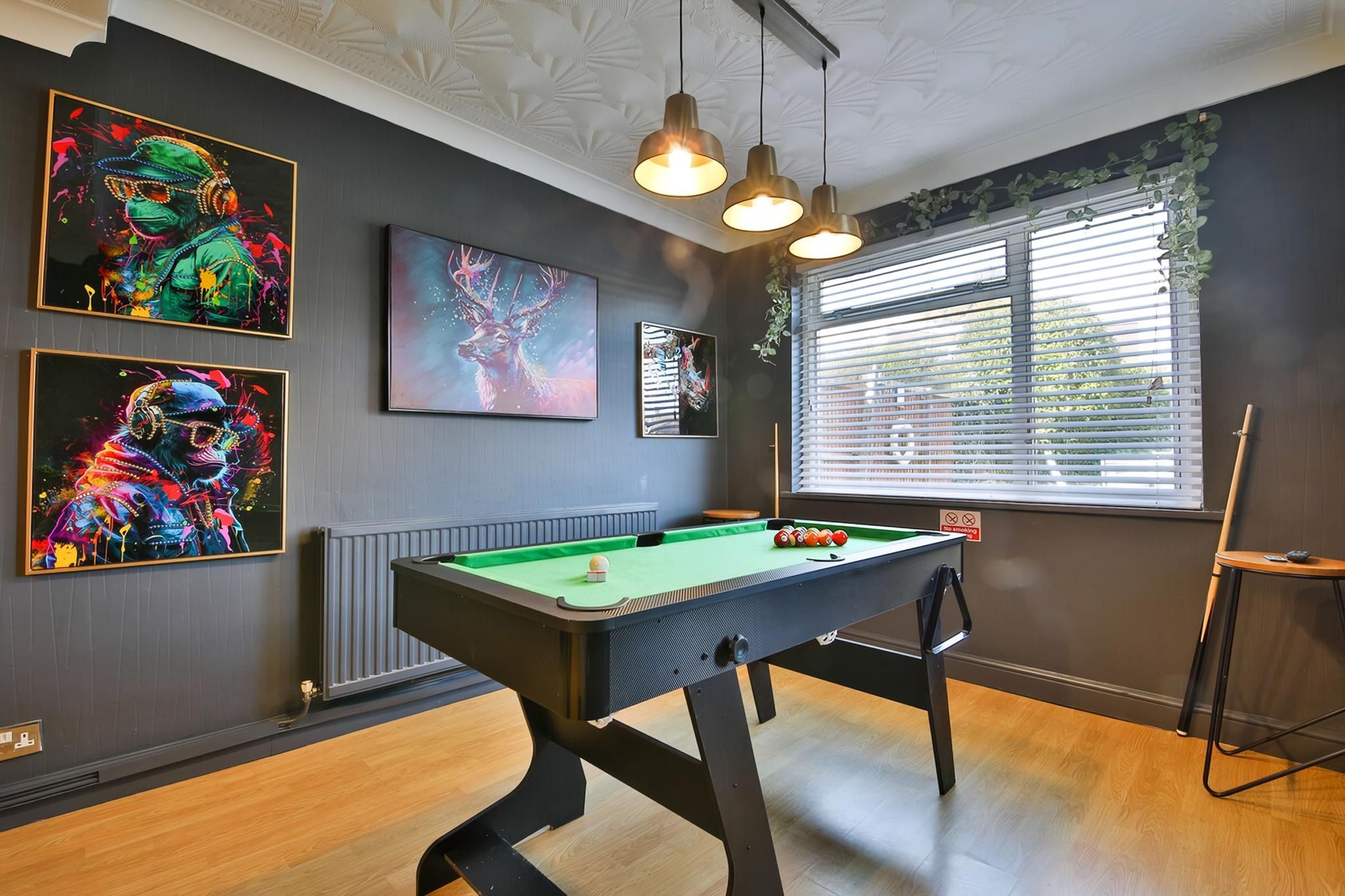 A pool table with a green felt surface, surrounded by walls with framed artwork, a window with blinds, and a ceiling with recessed lighting.