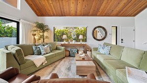 Una sala de estar moderna con techo de madera, sofás verdes, una mesa de centro y un ventanal con persianas.