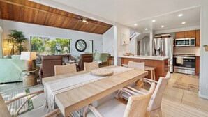 Una cocina moderna con electrodomésticos de acero inoxidable, un comedor con una mesa de madera y sillas de mimbre, y una sala de estar con un sofá verde y una planta en maceta.