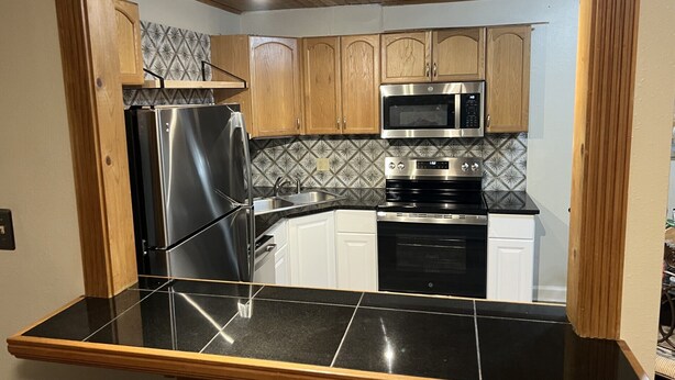 A modern kitchen with a black countertop, stainless steel appliances, wooden cabinets, and a patterned backsplash. - Complete Remodel 1 Block From the River Center, Minutes to LSU (Baton Rouge)