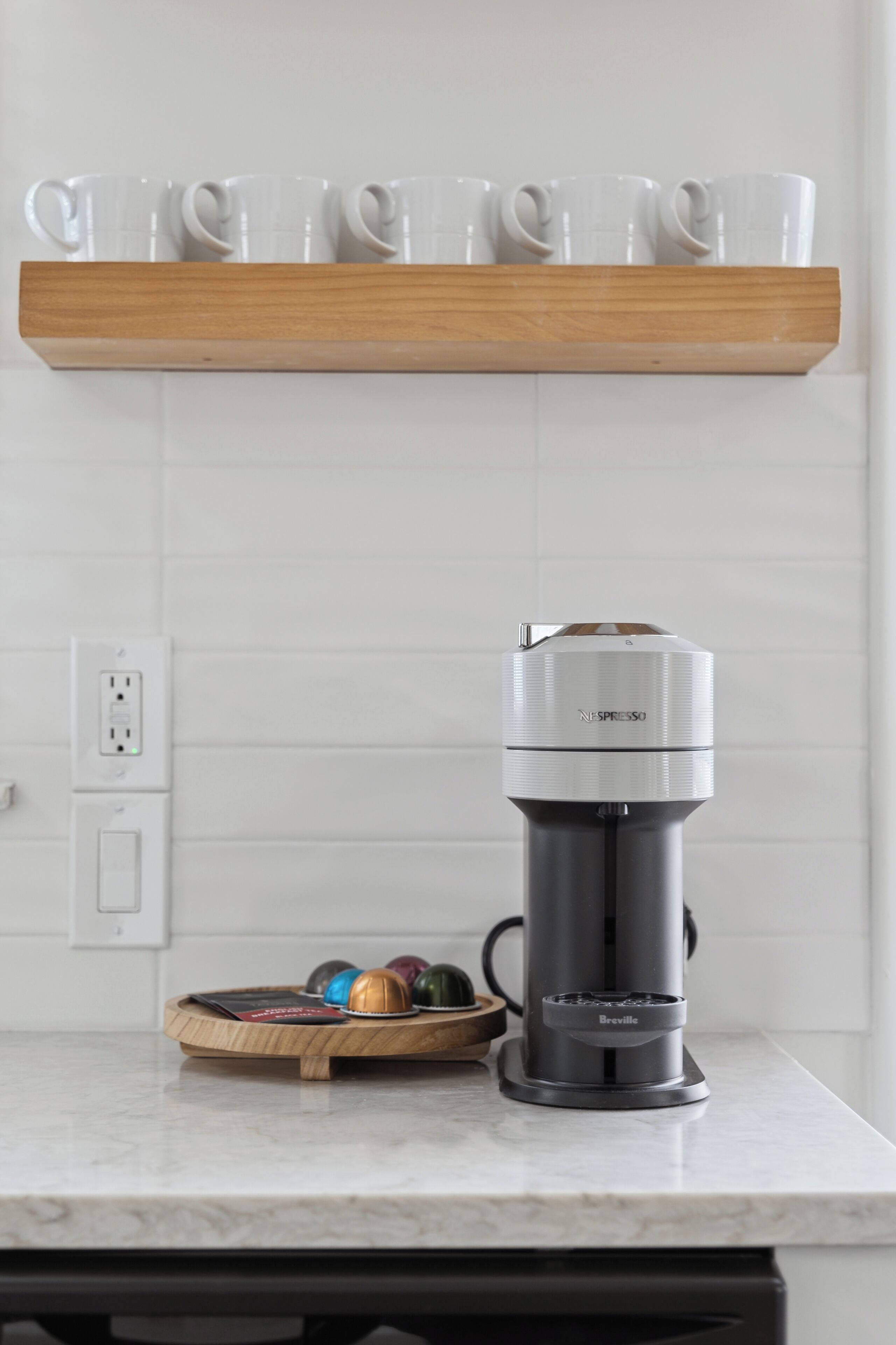 A kitchen with a coffee machine, a wooden shelf with cups, and a tray with coffee pods.