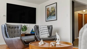 Un salón con dos sillones de cuero, una mesa de centro redonda de madera, un televisor de pantalla plana montado en la pared y un cuadro enmarcado sobre el televisor.