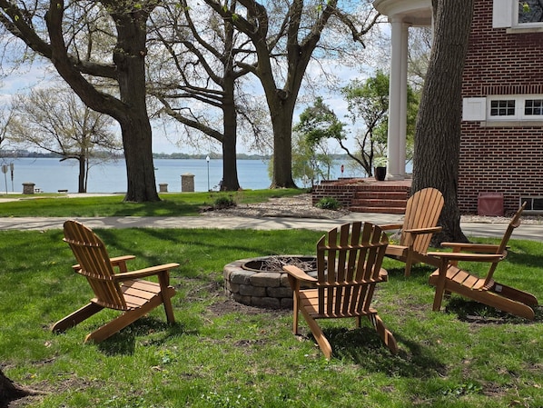 A gazebo with Adirondack chairs around a fire pit, overlooking a lake.