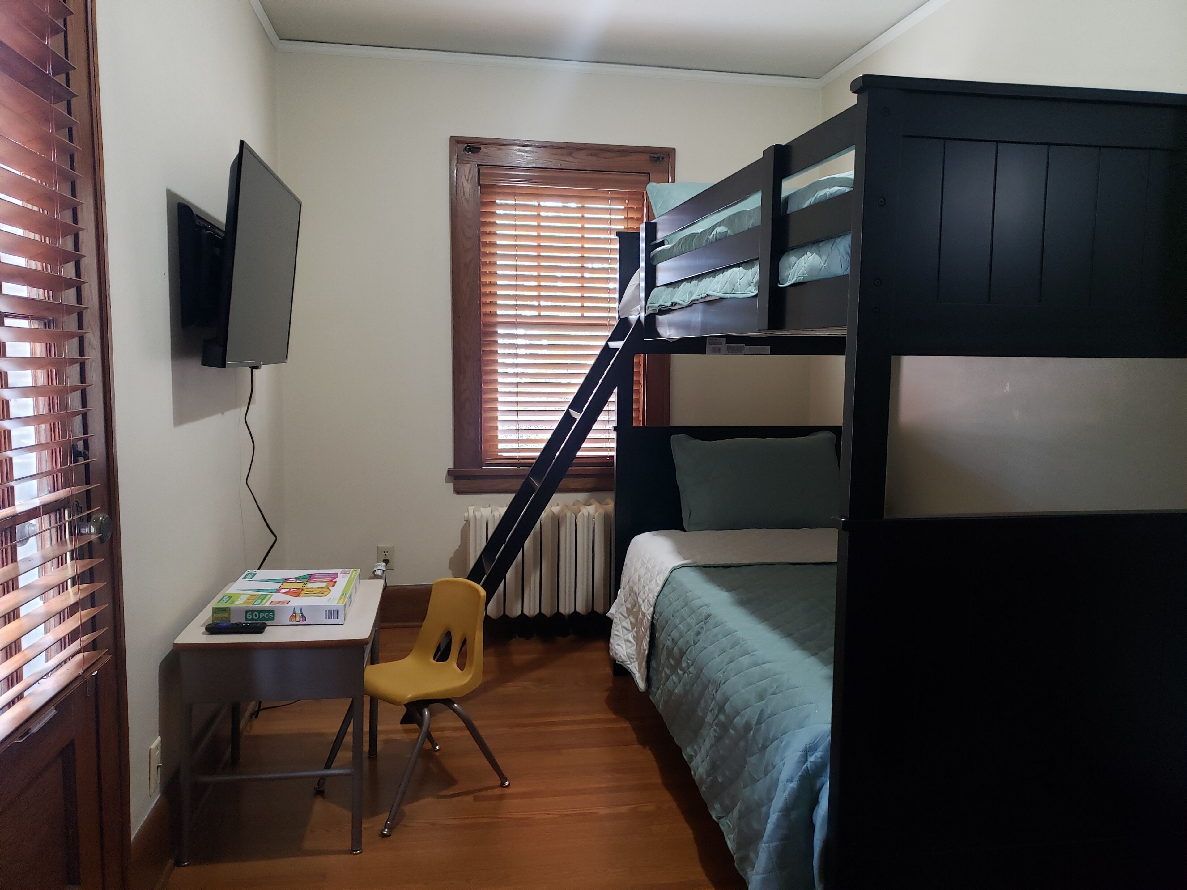 Ein Schlafzimmer mit Etagenbett, Schreibtisch mit Stuhl, Wandfernseher und Fenster mit Jalousien.