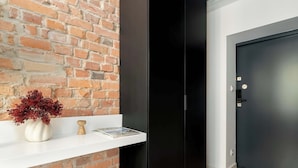 Une pièce avec un mur en briques, une étagère blanche ornée d’un vase et d’un livre, une armoire noire et une porte sombre.