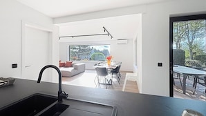 Una cocina moderna con encimera y lavamanos negros, un comedor con mesa y sillas, y vistas al exterior a través de grandes ventanales.
