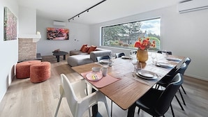 Un moderno comedor con una mesa de madera dispuesta para cuatro personas, rodeada de sillas negras. Hay un sofá con cojines naranjas y una mesa baja con un posavasos rosa. Un ventanal ofrece vistas a árboles y un edificio.