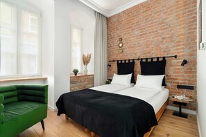 A bedroom with a brick wall, a bed with black and white bedding, a green chair, and a small bedside table with a lamp.