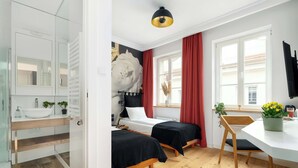 Una habitación con dos camas, un lavamanos blanco, un espejo, una ventana con cortinas rojas y suelo de madera.