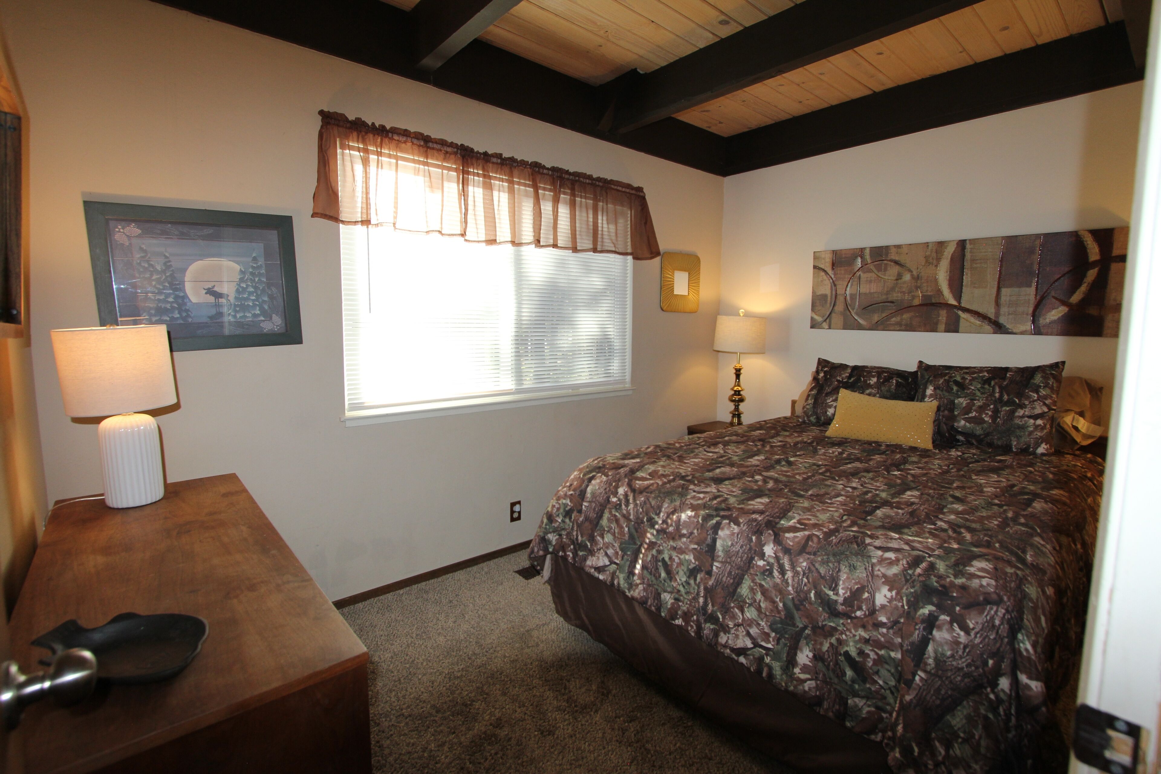 A bedroom with a large bed, two bedside lamps, a framed picture on the wall, and a window with blinds.