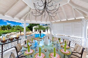 A covered outdoor dining area with a glass table, chairs, and a pool in the background. - Sugar Hill - Half Century House 6 Bedroom Villa (Holetown)