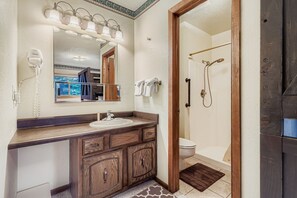 A bathroom with a wooden vanity, a sink, a mirror with lights, a hairdryer, and a shower area. - Snowdance C304 2 Bedroom Condo (Keystone)