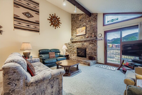 A cozy living room with a stone fireplace, leather sofas, and a large window offering a scenic view. - Snowdance C304 2 Bedroom Condo (Keystone)