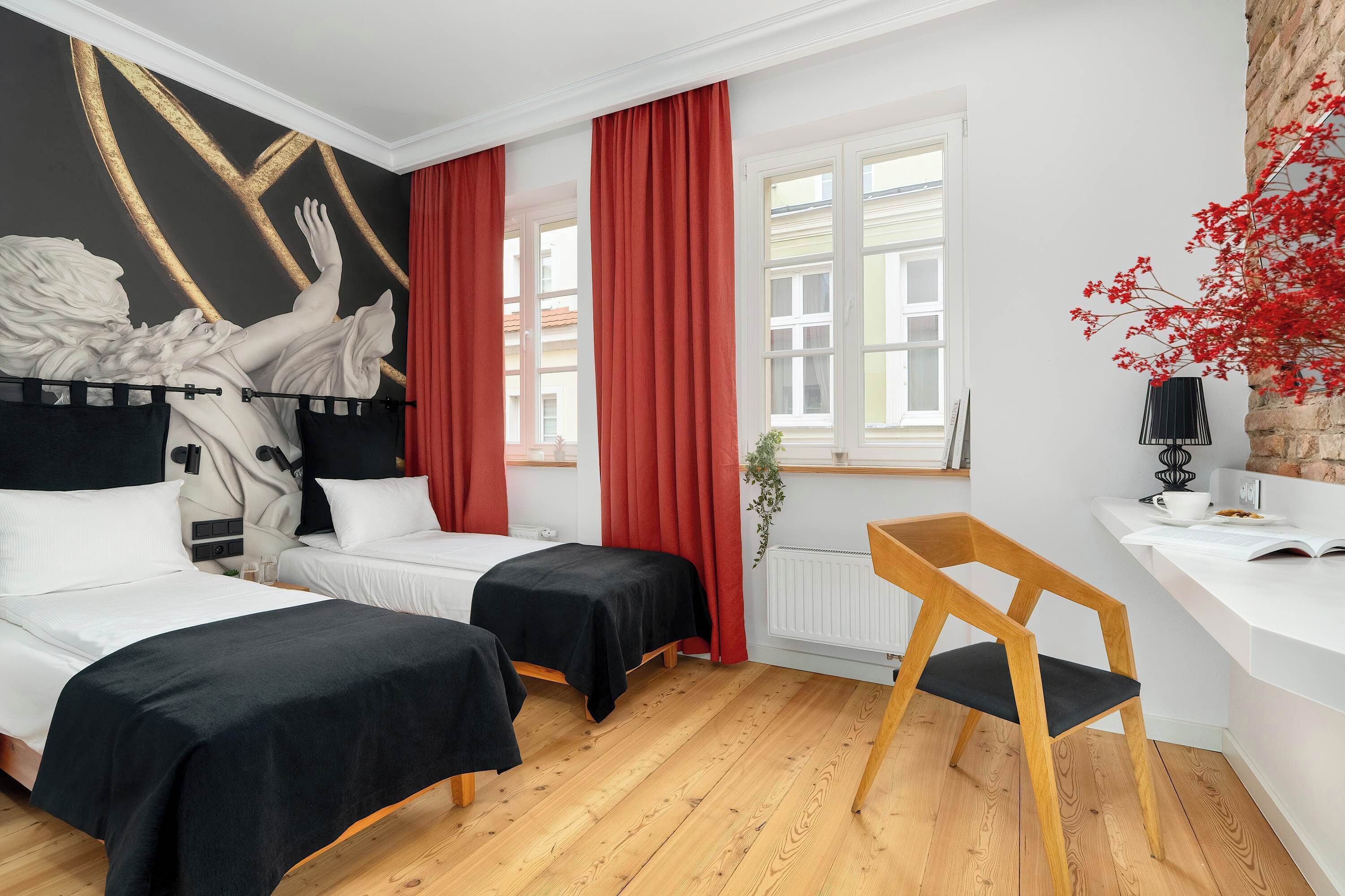 A hotel room with two beds, a wooden floor, a red curtain, a white wall with a gold design, and a window with red curtains.