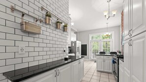 A kitchen with white cabinets, black countertops, and a tiled backsplash. There are hanging plants, a window with a view, and a chandelier. - High Street Hideaway - For 8 (Columbus)