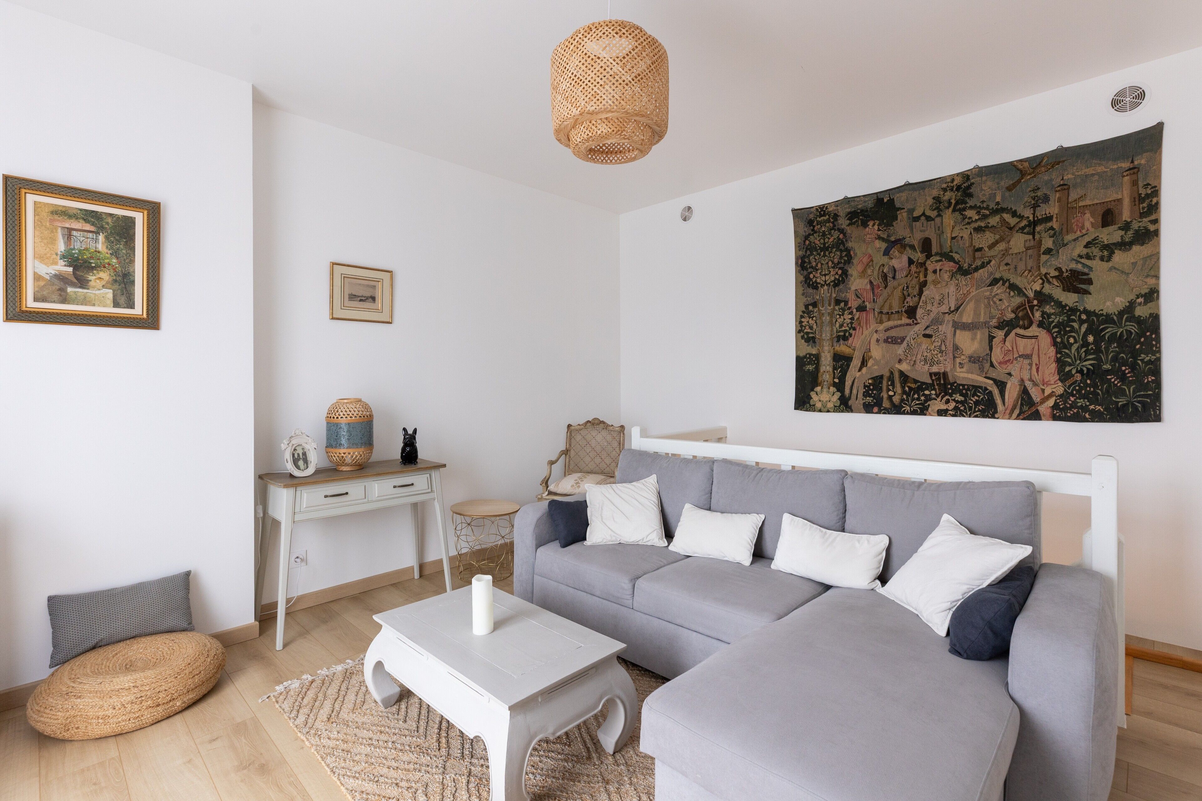 Un soggiorno moderno con un divano angolare grigio, un tavolino da caffè bianco, un pouf in vimini, una piccola scrivania bianca e un grande arazzo appeso al muro.