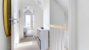 Un corridoio stretto con una porta bianca che dà su una camera da letto. Un piccolo armadietto bianco è appoggiato al muro, e a destra si intravede una panca di legno.