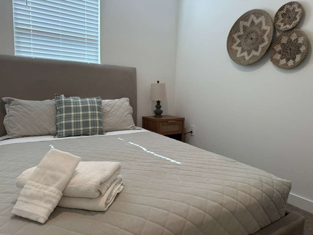 Een net opgemaakt bed met een beige deken, een geruit kussen en een opgevouwen handdoek ernaast. Er staat een nachtkastje met een lamp en een decoratief bord aan de muur.