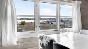Huoneessa on suuri ikkuna, valkoiset verhot, valkoinen pöytä ja pehmeä huopa tuolilla.
