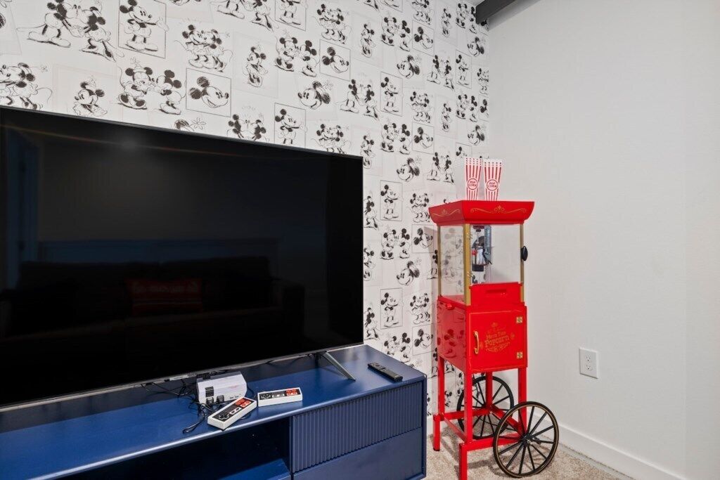 Une pièce avec un grand téléviseur à écran plat posé sur un meuble bleu, un chariot rouge avec un plateau rouge, et un mur recouvert d’un papier peint représentant des personnages de dessins animés.