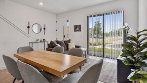 Una sala de estar moderna con una mesa de comedor de madera, sillas tapizadas en gris, un sofá con cojines, un ventanal con persianas y una planta en maceta.