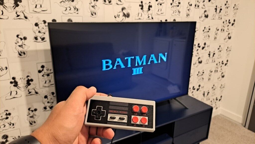 Un televisor mostra 'BATMAN III' i una mà sosté un comandament de joc al davant.