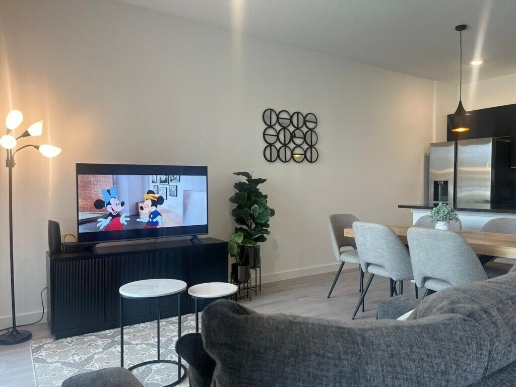 A modern living room with a flat-screen TV, grey sofas, a coffee table, a dining area with chairs, and a decorative wall piece.
