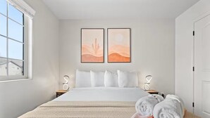 Ein modernes Schlafzimmer mit einem großen Bett, zwei an der Wand befestigten Kunstwerken, einem Fenster mit Blick und einem ordentlich bezogenen Bett mit Handtüchern.