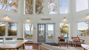 A spacious room with large windows, a wooden table, chairs, and a bench. - Owl's Landing: Bright 3-Bed Coastal Home with Loft, Screen Room & Ocean Views (Prince Edward Island)