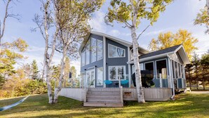 A modern two-story house with large windows, a deck, and a covered outdoor seating area. - Owl's Landing: Bright 3-Bed Coastal Home with Loft, Screen Room & Ocean Views (Prince Edward Island)