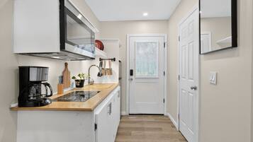 A narrow hallway leading to a white door, with a kitchen area on the left featuring a stove, coffee maker, and cabinets.