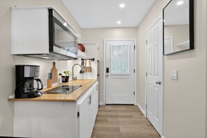 A narrow hallway leading to a white door, with a kitchen area on the left featuring a stove, coffee maker, and cabinets.