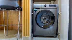 Lavadora compacta y moderna con pantalla digital, ubicada en un pequeño cuarto de lavado con paneles de madera y una silla de metal.