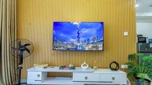 Una sala de estar moderna con una televisión de pantalla plana montada en una pared de madera, un mueble blanco para entretenimiento con libros y objetos decorativos, un ventilador de pie y una planta en maceta.