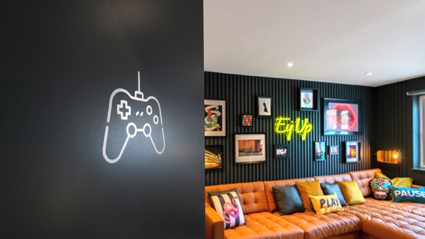 A modern living room with a large orange sectional sofa, a neon sign that reads "Play," and a wall-mounted game controller graphic. - Peveril House  (Nottingham)
